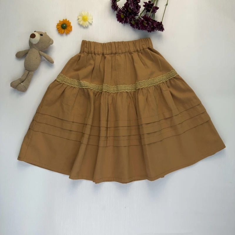 دامن دخترانه کتان رنگ شتری با تزیینات توری و چین‌دار روی زمینه سفید.